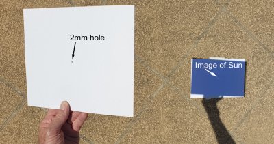 The photo shows a hand holding a card with a 2mm hole in it. A small and quite faint image of the Sun is projected onto a white paper about one metre away.