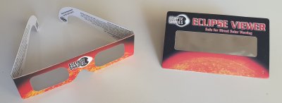 The photo shows a pair of eclipse glasses on the left as a cardboard frame that can be worn similar to spectacles but with solar filter material where the lenses would be. On the right is a solar viewer as a flat card with filter material in its central section such that it can be held in front of the face and the Sun viewed through the filter with both eyes